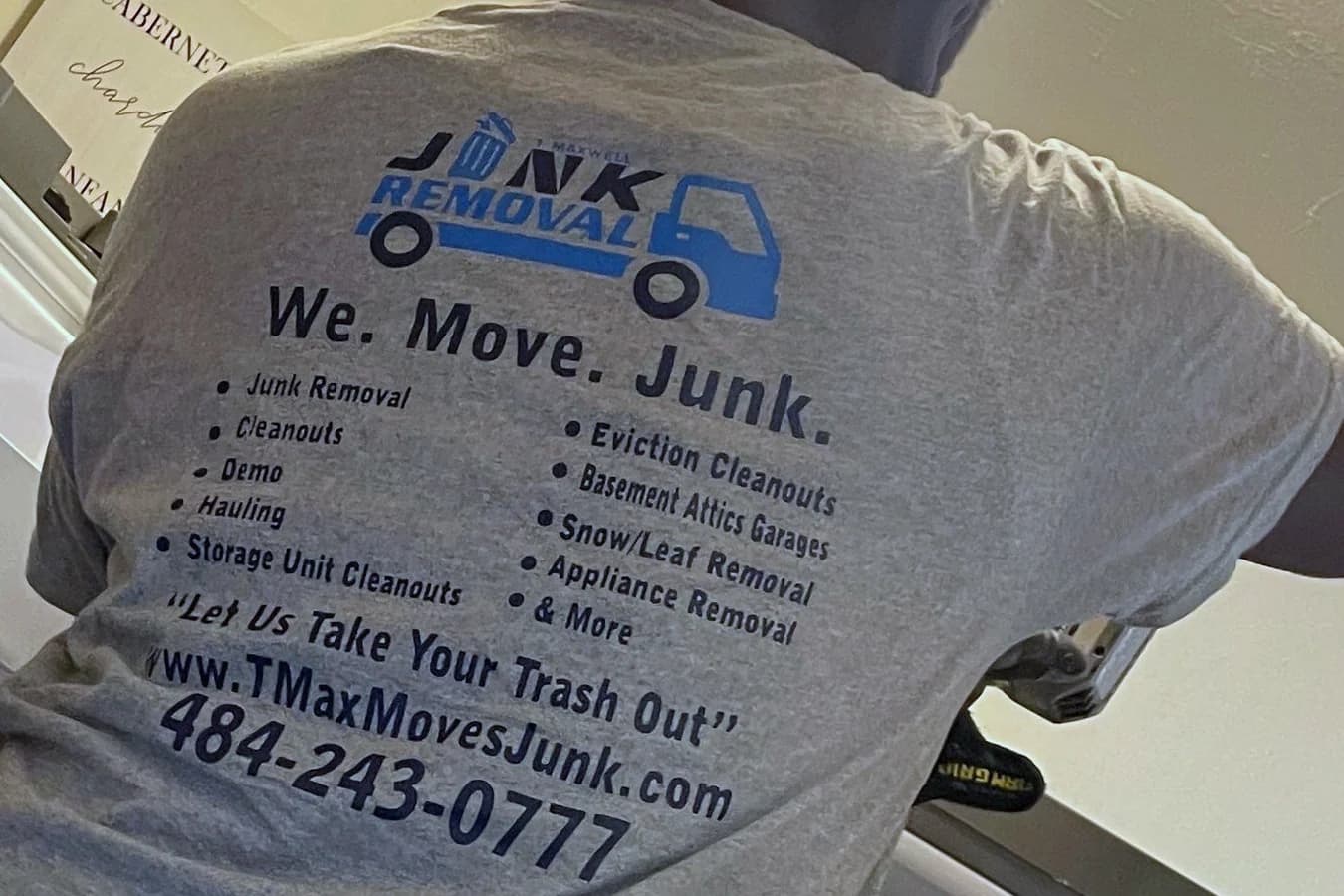 Close view of a T Maxwell crew member's grey work shirt, printed with the company's junk removal truck logo, a list of services, the website, and the phone number.