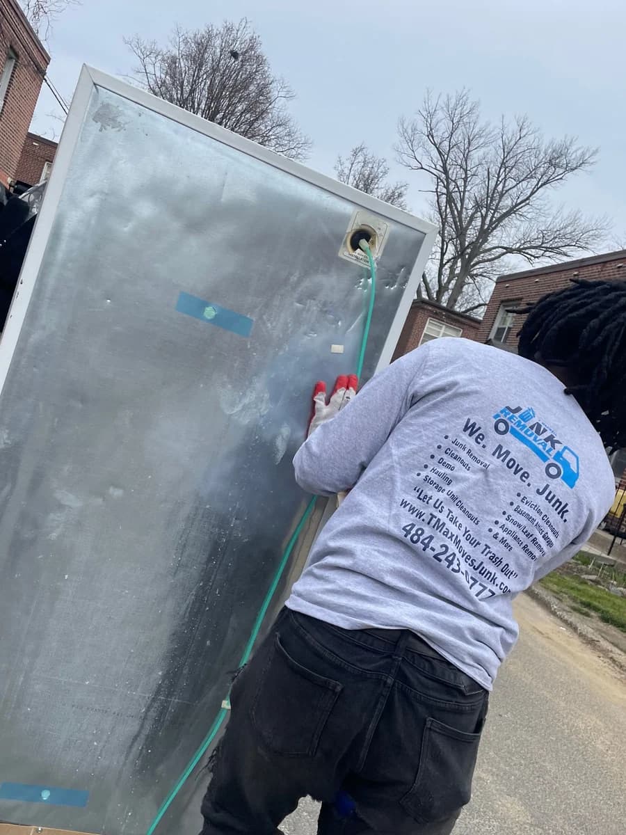A T Maxwell crew member in work gloves walks a full-size refrigerator away from a rowhome toward the dump trailer parked at the curb.